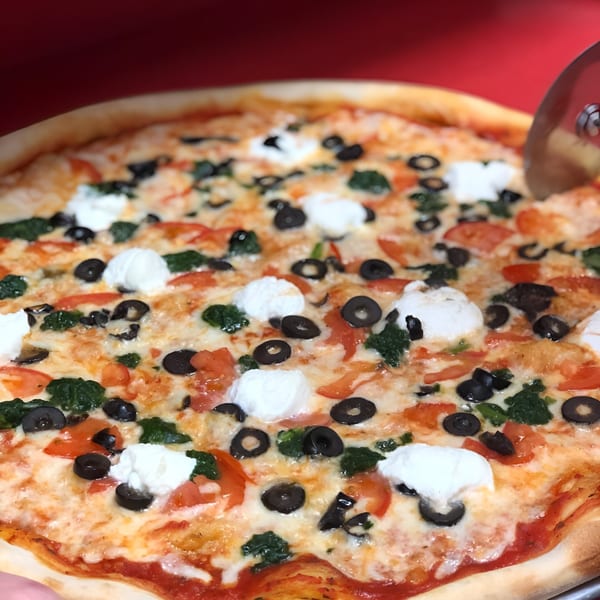 King's Pizza with black olives, tomatoes, spinach, and dollops of ricotta cheese being sliced.